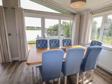 A dining room with a wooden table and six blue upholstered chairs next to large windows overlooking a lake at 40 Delamere Point in Delamere