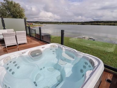 An outdoor hot tub on a wooden deck overlooking a lake with patio chairs and a glass railing at 40 Delamere Point in Delamere