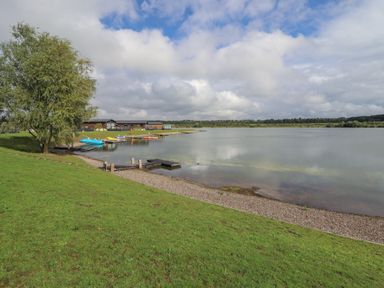 A lakeside with boats docked near a grassy shore and buildings in the background at 40 Delamere Point in Delamere
