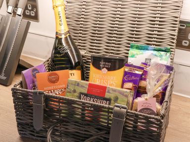 A wicker basket on a kitchen counter containing a bottle of Canti prosecco, Yorkshire crisps, Guppy's chocolates, Cadbury drinking chocolate sachets, Yorkshire tea, and a packet of Sawley Kitchen sweets