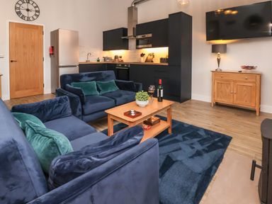 A living area with blue sofas green cushions a wooden coffee table kitchen cabinets a refrigerator a wall clock a wooden sideboard and a wall mounted television at Robins Nest in Pocklington