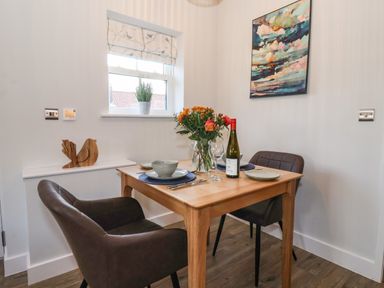A small dining area with a wooden table set for two with a wine bottle flowers and tableware and two chairs by a window with a plant and a wall painting at Robins Nest in Pocklington