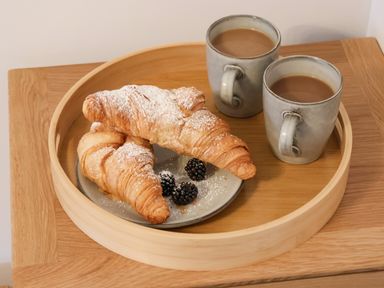 A wooden tray with two croissants dusted with powdered sugar and three blackberries on a plate alongside two mugs of coffee