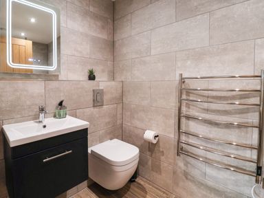 A bathroom with a wall-mounted sink, a wall-hung toilet, a rectangular mirror with lights, and a towel radiator at Robins Nest in Pocklington