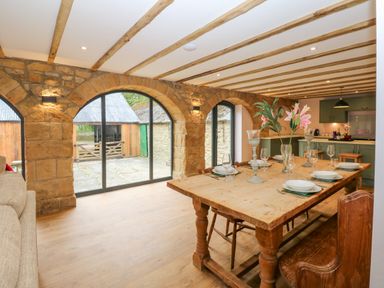 A dining area with a wooden table set for six placed near arched stone windows and a kitchen in the background at The Coach House in Great Whittington near Corbridge