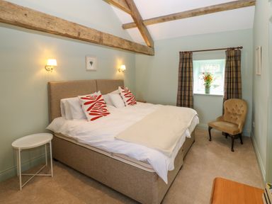 A bedroom with a double bed two red and white cushions a beige chair a small white side table and a window with plaid curtains at The Coach House in Great Whittington near Corbridge