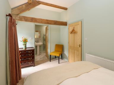 A bedroom with a bed a yellow chair a wooden dresser with flowers and an open door to a bathroom at The Coach House Great Whittington near Corbridge