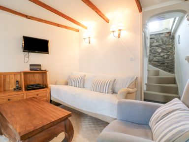 A living room with a wooden table sofa and armchair a wooden cabinet with a television on the wall and stairs with a stone wall at Aunt Dora's in Port Isaac