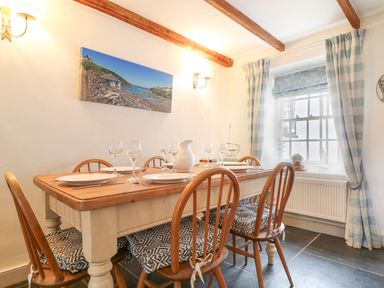 A dining room with a wooden table set with plates glasses and a pitcher surrounded by wooden chairs with cushions and a window with curtains at Aunt Doras in Port Isaac