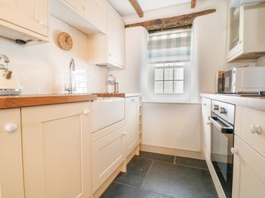 A kitchen with wooden countertops white cabinets a sink an oven a microwave a window with a checkered blind and exposed wooden ceiling beams at Aunt Doras in Port Isaac