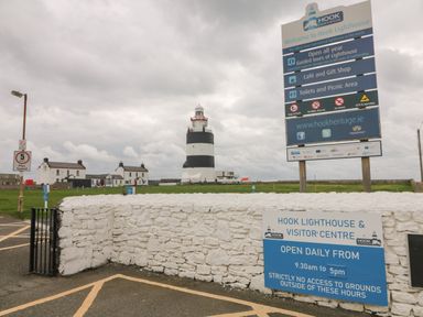 A lighthouse behind a stone wall with signs about visiting hours and rules at Hook Lighthouse Slade near Fethard-On-Sea County Wexford