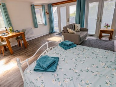 A room with a bed covered with floral sheets and towels a gray sofa wooden dining table with chairs and glass doors with blue curtains at Tower Cottage Near Pembroke