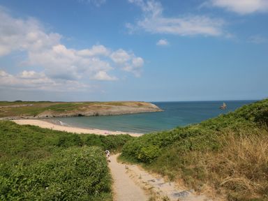 A pathway leading to a sandy beach with people near a bay surrounded by grassy hills and a rock formation in the sea
