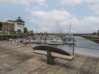 A bench shaped like a whale tail on a stone path next to a marina with boats and a building labeled beacon in Whitehaven
