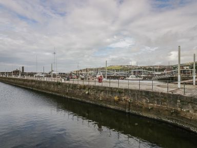 A harbor with boats docked by a stone quay and a curved pedestrian bridge with hills in the background at 8 Corkickle in Whitehaven