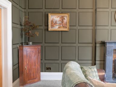 A corner of a room with green paneled walls a wooden cabinet with a plant on top a painting hanging on the wall a gray carpet brown leather sofa with cushions and a black fireplace at 8 Corkickle in Whitehaven