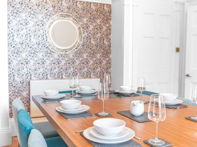 A dining room with a wooden table set with white dishes and wine glasses and blue chairs at 8 Corkickle in Whitehaven