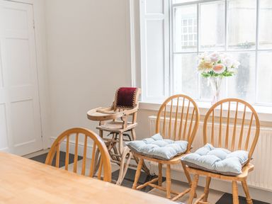 A dining area with two wooden chairs with cushions a wooden high chair and a vase of flowers on the window sill at 8 Corkickle in Whitehaven