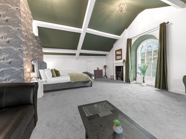 A bedroom with a bed bedside tables an armchair a fireplace and a large arched window with curtains at 8 Corkickle in Whitehaven