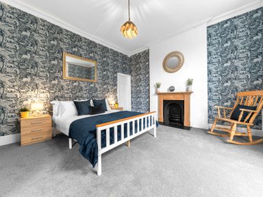 A bedroom with a white bed frame wooden rocking chair fireplace and patterned wallpaper at 8 Corkickle in Whitehaven