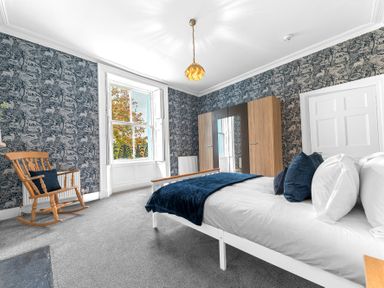 A bedroom with a bed and pillows a wooden rocking chair a wardrobe and a large window at 8 Corkickle in Whitehaven