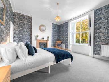 A bedroom with a white bed and blue pillows and blanket a wooden chair a fireplace and a large window at 8 Corkickle in Whitehaven