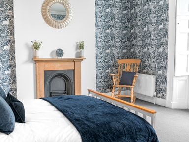 A bedroom with a bed covered with blue blankets a wooden armchair with a blue cushion and a fireplace with decorative vases and mirrors at 8 Corkickle in Whitehaven