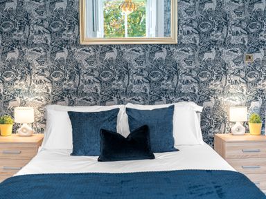 A bedroom with a bed with blue pillows and throw a wall with animal patterned wallpaper and two wooden bedside tables each with a lamp and small plant at 8 Corkickle in Whitehaven
