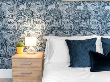 A bedroom with a wooden bedside table with a lamp and plant and a bed with white linens and dark blue pillows at 8 Corkickle in Whitehaven