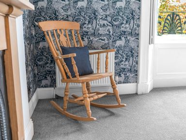 A wooden rocking chair with a dark cushion on a carpeted floor against a wall with animal patterned wallpaper at 8 Corkickle in Whitehaven