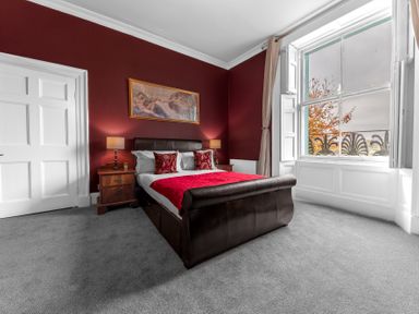 A bedroom with a dark leather bed red pillows and blanket two bedside tables with lamps and a large window at 8 Corkickle in Whitehaven