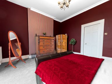 A bedroom with a wooden dresser, wardrobe, standing mirror, red bedspread, and maroon walls at 8 Corkickle in Whitehaven