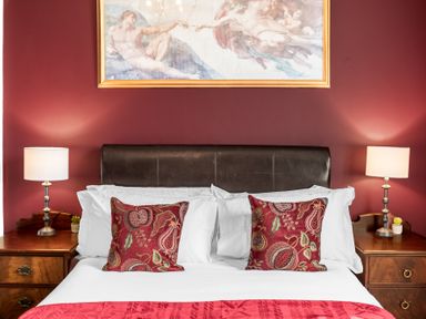 A bed with white sheets and red patterned pillows against a red wall with a framed artwork above and bedside tables with lamps at 8 Corkickle in Whitehaven