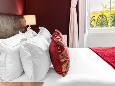 A bed with white pillows and red decorative cushions beside a window with white curtains at 8 Corkickle in Whitehaven