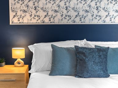 A bed with white sheets and three blue pillows next to a wooden nightstand with a lamp and small plant at 8 Corkickle in Whitehaven