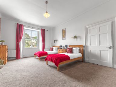 A bedroom with two single beds with red blankets and pillows a window with red curtains a wooden dresser and a door in 8 Corkickle Whitehaven