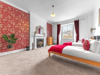 A bedroom with two single beds with red throws and pillows a fireplace a wooden chest of drawers and red patterned wallpaper at 8 Corkickle in Whitehaven