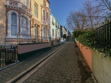 A narrow paved street lined with multicolored houses on the left and a green hedge with a fence on the right under a clear blue sky at 8 Corkickle in Whitehaven