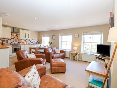 An open-plan living room with brown leather chairs and sofa a kitchen with white cabinets and tiled backsplash and a dining table at Crab Corner in Wells-Next-The-Sea