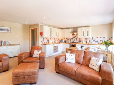An open plan living area with brown leather sofas and armchair a kitchen with white cabinets and a wooden dining table with breakfast items at Crab Corner in Wells-Next-The-Sea