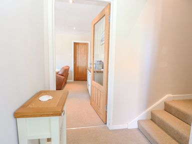 A hallway with stairs carpeted in beige and a small wooden table with a shell on it leading to a room with a brown sofa at Crab Corner in Wells-Next-The-Sea