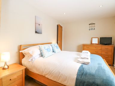 A bedroom with a wooden bed with white bedding and blue crab pillows beside a wooden nightstand with a lamp and a wooden dresser with a TV and mirror at Crab Corner in Wells-Next-The-Sea