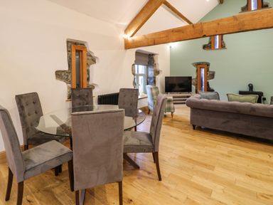 A dining area with a glass round table and six upholstered chairs next to a living room with two sofas and a television at The Barn by Lyons in Talybont near Barmouth
