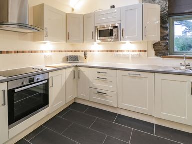 A kitchen with white cabinets black floor tiles built in oven microwave and window at The Barn by Lyons Talybont near Barmouth