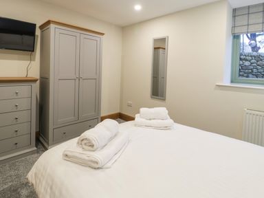 A bedroom with a bed with towels on it a wardrobe a chest of drawers a wall mounted TV a window and a wall mirror at The Barn by Lyons in Talybont near Barmouth