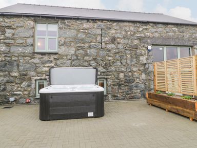 An outdoor hot tub on a paved area next to a stone wall with windows and a wooden planter at The Barn by Lyons in Talybont near Barmouth