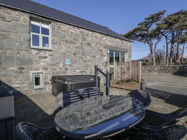 An outdoor patio area with a stone wall building a glass table and black wicker chairs and a hot tub at The Barn by Lyons in Talybont near Barmouth