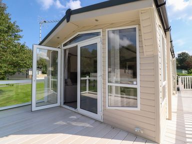 An exterior view of a light beige mobile home with white framed glass doors and windows on a wooden deck at 7 The Oaks in Long Preston near Hellifield