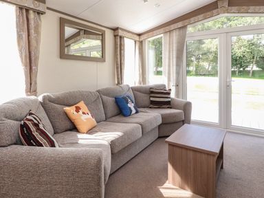 A living room with a grey corner sofa with patterned and dog-themed cushions a wooden coffee table and large windows at 7 The Oaks Long Preston near Hellifield