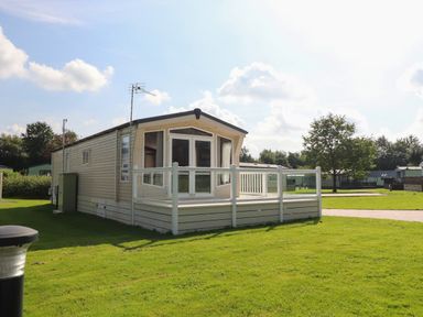 A mobile home with a small fenced deck on a grassy plot with trees and other homes in the background at 7 The Oaks in Long Preston near Hellifield